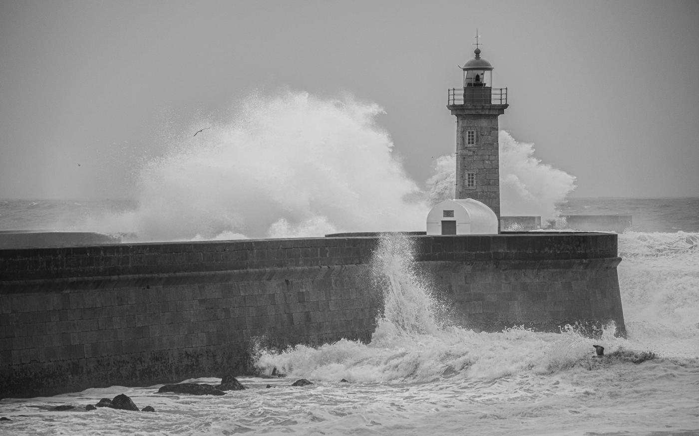 a black and white photo of waves crashing against a lighthouse by Gary Walker-Jones courtesy of Unsplash.