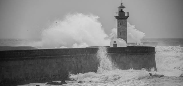 a black and white photo of waves crashing against a lighthouse by Gary Walker-Jones courtesy of Unsplash.